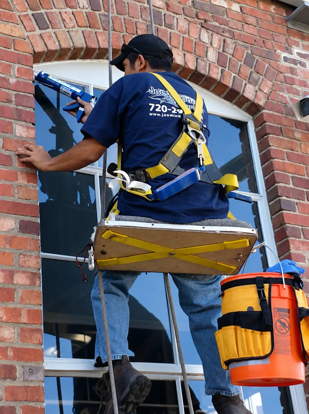 Joe's Window Cleaning technician on rope access applying caulk to a brick building window