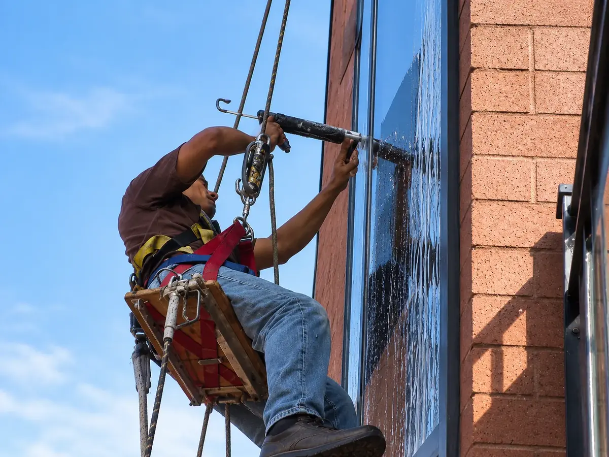Joe's Window Cleaning technician on rope access applying caulk to a commercial storefront window