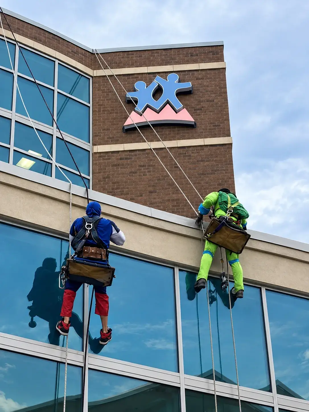 Two rope access technicians in superhero costumes cleaning the glass of a brick building