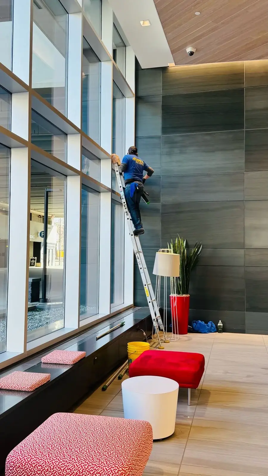Joe's Window Cleaning technician on a ladder cleaning floor-to-ceiling glass in a building lobby